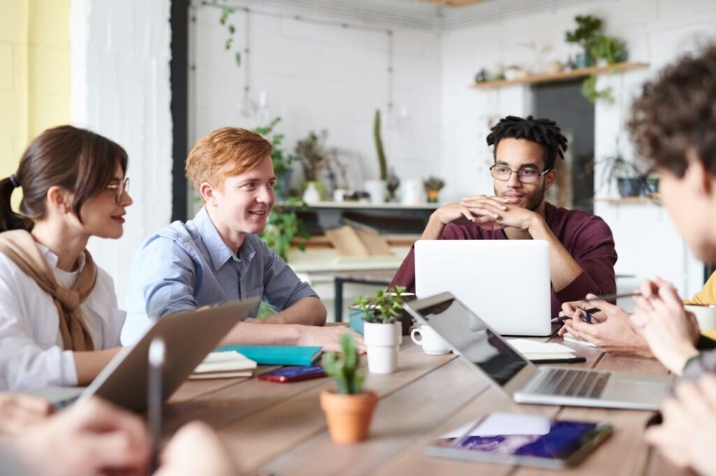 A diverse group of adults collaborating in a modern office setting with laptops and plants.